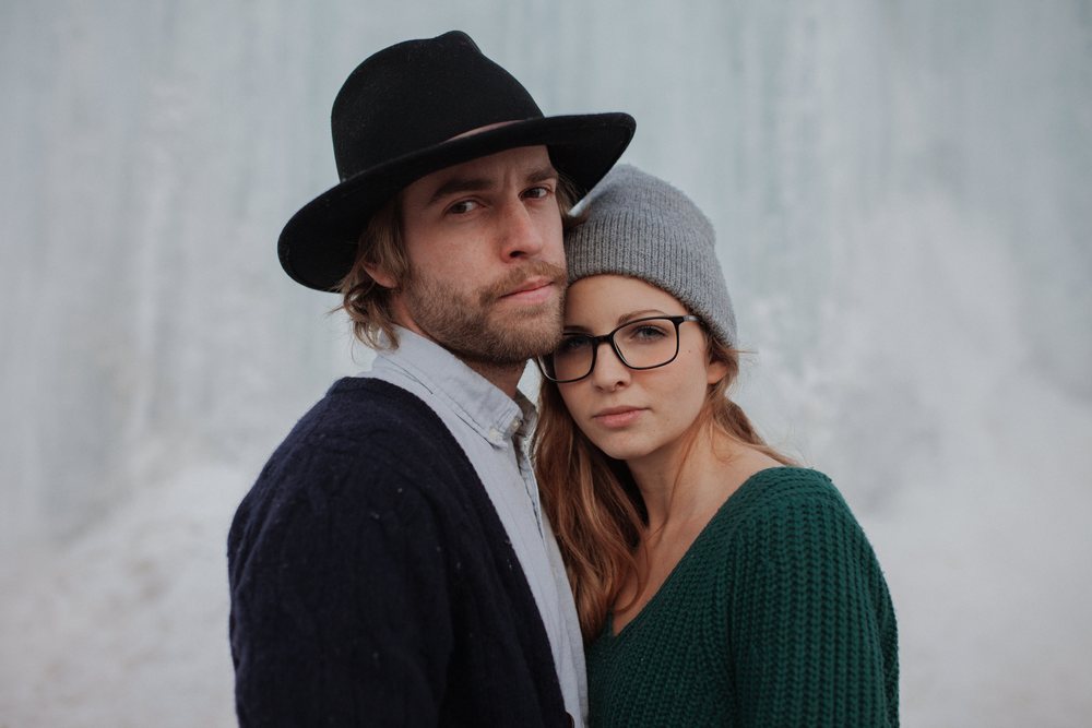 A couple bundled in winter hats and knits stands cheek to cheek against a pale wash of ice and snow.