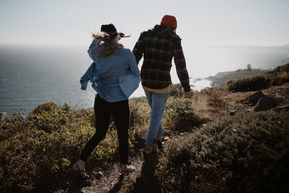 A couple walks away along a narrow path on a coastal headland. The ocean below them and the wind pulling at their clothes.